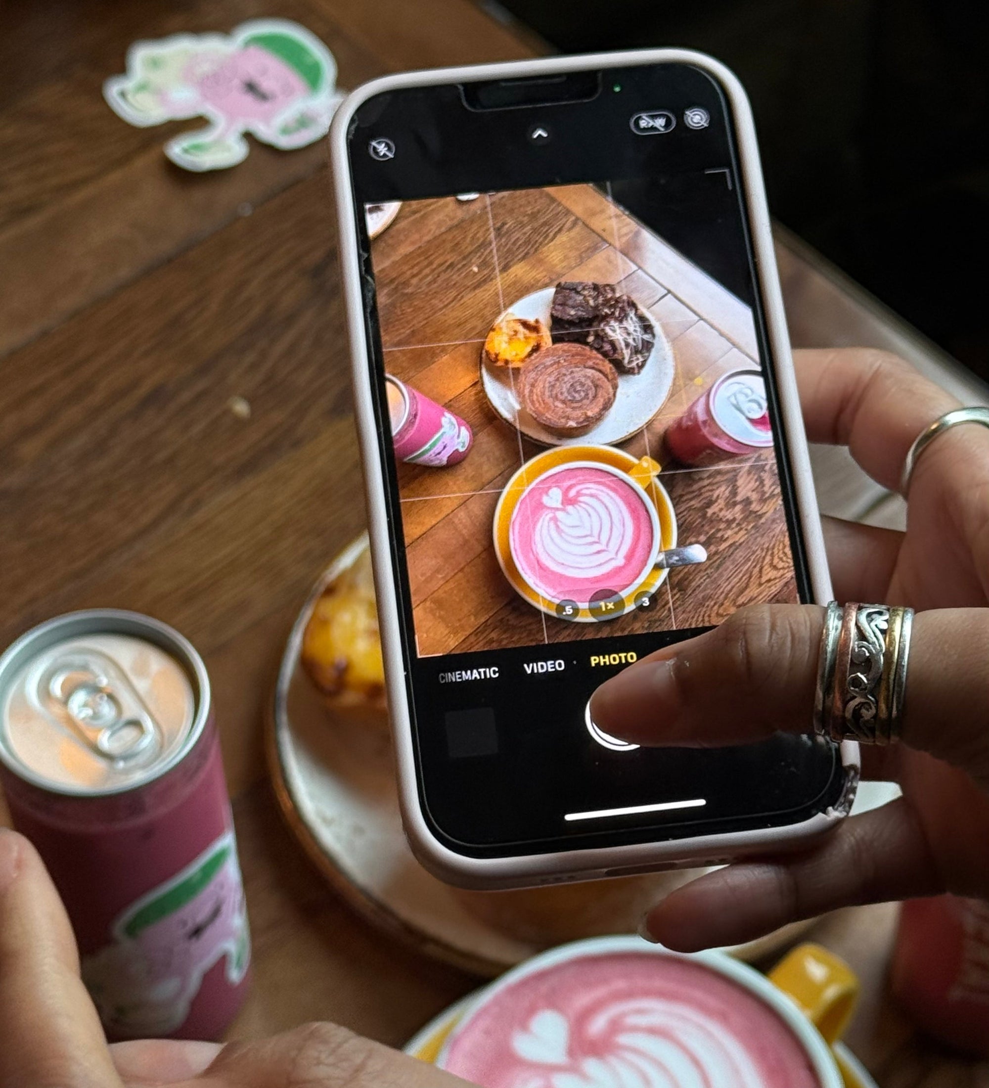 Person taking a photo of a Blendsmiths Pink Chai latte with latte art, pastries and canned Pink Chai drinks on a café table.