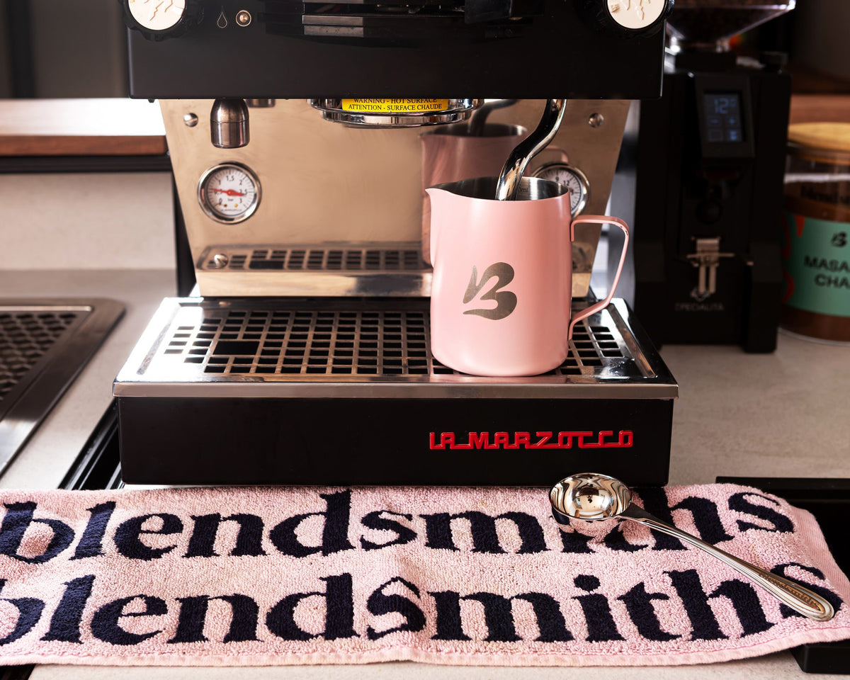 A La Marzocco espresso machine with a Blendsmiths pink milk pitcher resting on its drip tray, set in a cafe  environment with a Blendsmiths barista towel in the foreground.
